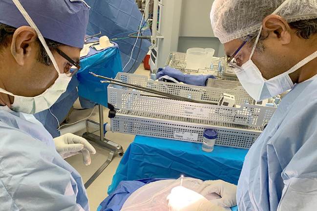 Two surgeons in full operating gear examine a specimen in an iced bowl.