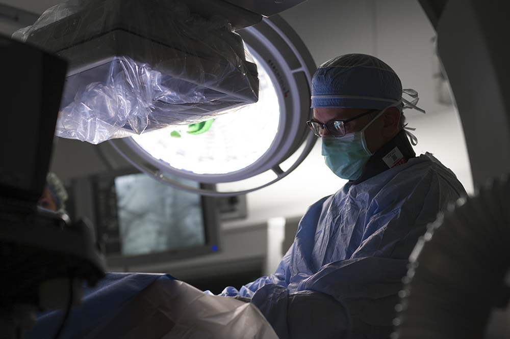 Pediatric cardiologist Dr. John Rhodes at the operating table. He is wearing a cap and surgical gown. There is a large light to the left.